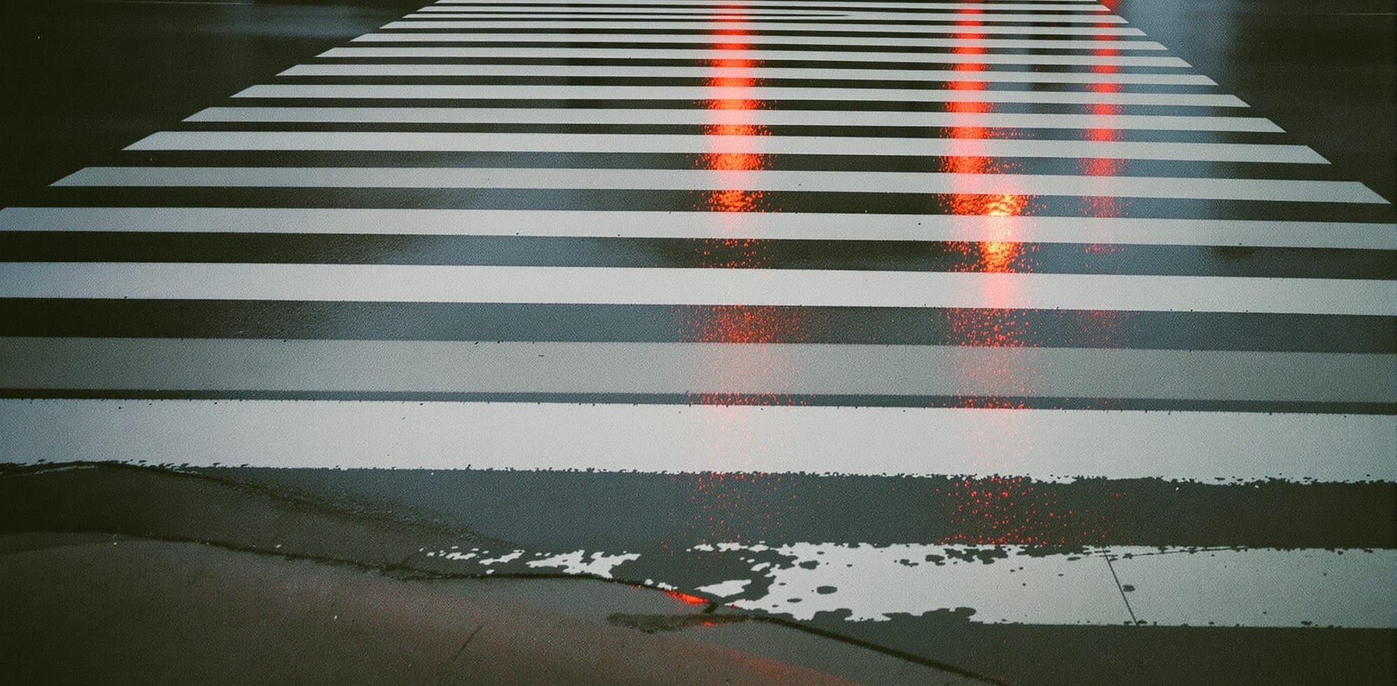 Rain-wet crosswalk stripes at dusk with red signal light reflected on the asphalt