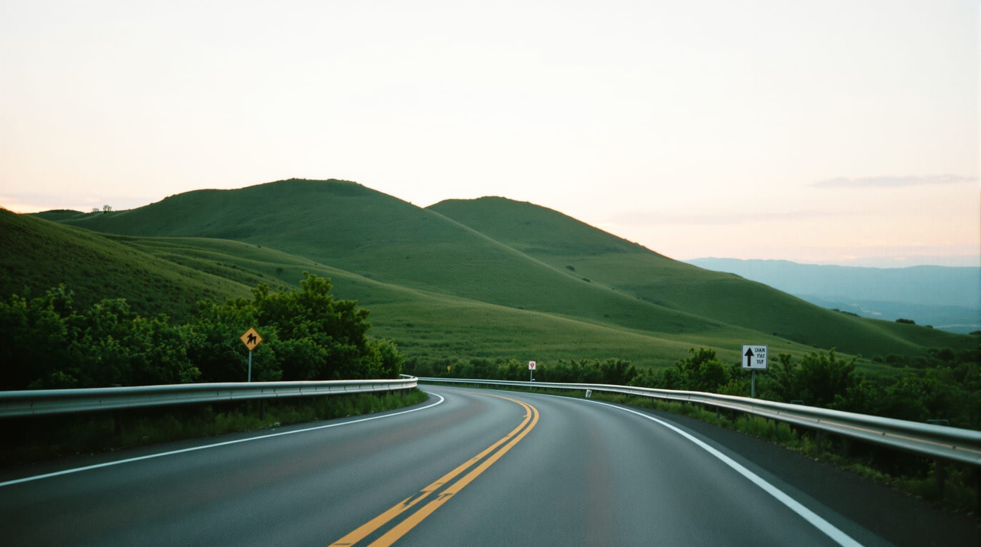 Two-lane road curving through green hills at dusk with a double yellow line and guardrail